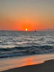 A breathtaking sunset unfolds over the Pacific Ocean, as seen from Venice Beach, with the silhouette of sailboats and the distant mountains of Malibu creating a serene atmosphere.