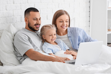 Cute little girl with her parents using laptop under blanket in bedroom