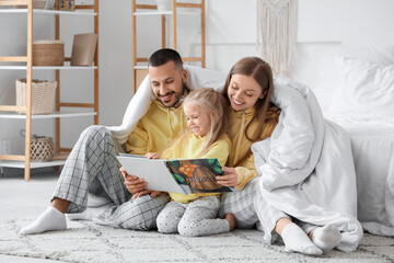 Happy parents with their little daughter reading book under blanket in bedroom