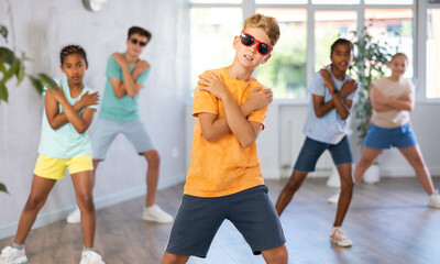 Obraz premium Boy in sun glasses performs choreographic exercises and teaches energetic mobile social dance Jazz-modern together with friends. Multi-racial group of children performs movements in spacious studio