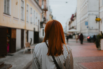 a woman with red hair walks along a street with bars and restaurants in a European city