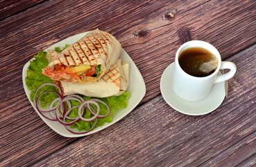 A cup of hot black tea from a bag and a plate with juicy meat shawarma on a lettuce leaf and chopped red onion on a dark wooden table.
