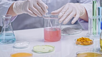 Experimental theme footage from close-up front view on white lab table with many petri dishes contain different experiment sample. Scientist stirring pink liquid and gray powder in glass beaker