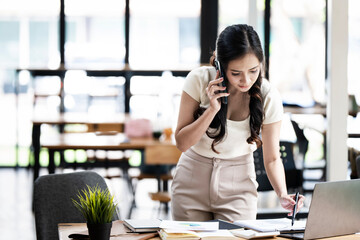 Cheerful business Asian woman freelancer making telephone call share good news about project working in office workplace, business finance concept.