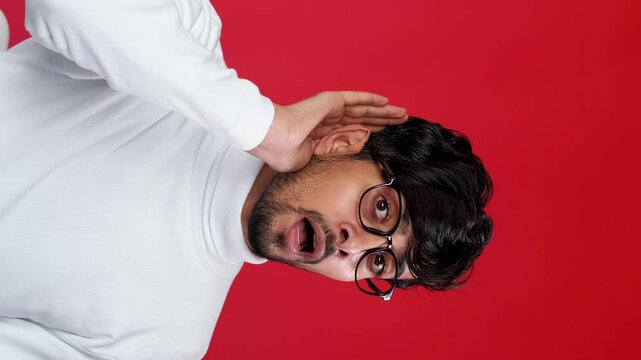 Vertical video. Gossip shock. Secret surprise. Curious stunned guy eavesdropping listening whisper news isolated on red copy space background.