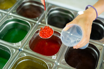 Hands of vendor preparing bubble tea, scooping red pearls from stainless steel container into...
