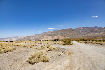 The ghost town of Ballarat, near Death Valley, California, sits abandoned in the desolate desert landscape, surrounded by rugged mountains and dry scrubland under a clear blue sky.