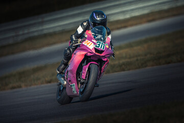  A person rides a pink motorcycle on a race track, surrounded by lush grass and a fence in the...