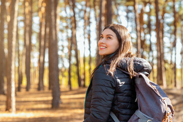 Pretty smiling girl with backpack enjoying view while walking by pine forest, copy space