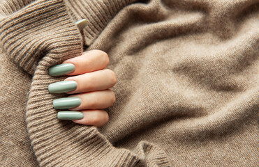 Elegant hand with long green nails holding a textured beige sweater during autumn fashion season