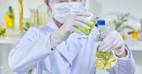 A scientist carefully pours a green liquid from a beaker into a flask containing a rosemary sprig and glass containers containing herbs in the backdrop. The picture show experiments in a lab setting.