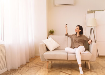 Split System. Satisfied european girl holding remote control relaxing under the air conditioner, sitting on couch