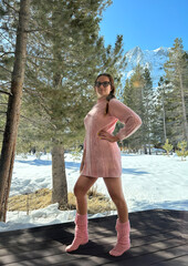 Woman in a pink outfit poses confidently amidst snow-covered scenery with mountains and pine trees in the background.