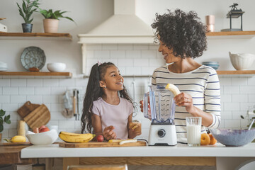 Smiling African American girl and her young mother making a banana smoothie on a large kitchen counter, enjoying fun and happy moments together in a cozy beige kitchen.
