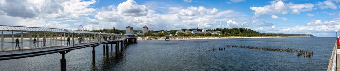 Stunning Panorama of a Coastal Landscape Featuring a Sandy Beach and Charming Small Town Viewed from the Pier, Under a Clear Blue Sky with Fluffy Clouds, Perfect for Summer Vibes