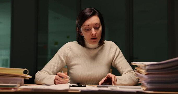 Woman works late at night in office, writes a report and looks at the time on smartphone
