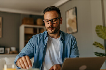 Fototapeta premium A focused mid aged male freelancer sitting at the wooden table at home and reading online projects on a laptop.