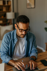 Fototapeta premium Handsome focused man in a denim shirt sitting at home and using a laptop