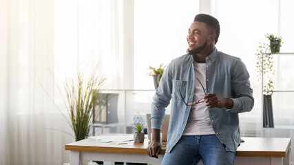 Work done. Happy african american startup owner leaning on office desk and looking aside, proud of...