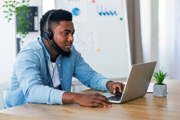 Young call center operator in headset solving issues with client online, using laptop at workplace in office
