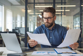 Fototapeta premium Surprised and serious young businessman working in the office, seated at a table and looking shocked at the documents and bills in his hands.