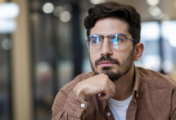 Obraz premium Pensive young man with glasses deep in thought about an idea indoors, seated in a bright and inviting space.
