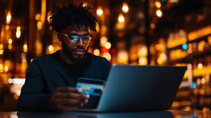 A young man uses a laptop while holding a credit card in a cozy, dimly lit cafe, showcasing modern online shopping.