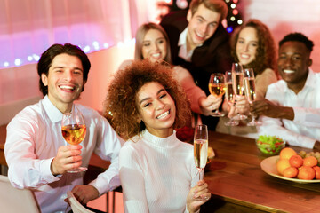 Cheerful Group Of Friends Having New Year Celebratory Dinner Holding Glasses Sitting Next To Christmas Tree At Home. Xmas Celebration