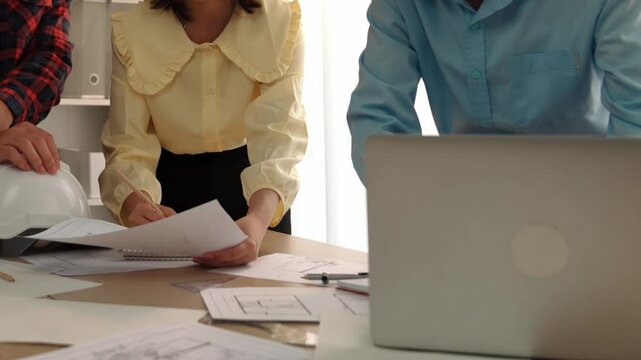 Architect and safety engineer discussion about building blueprint displaying on laptop on table with architectural document and wooden block scatter around at modern office. Closeup. Delineation.