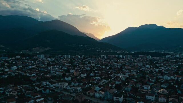 Aerial drone shot of Peja a city in Kosovo with a mountain in the background. The sun is setting and the sky is orange. The city is full of houses and buildings