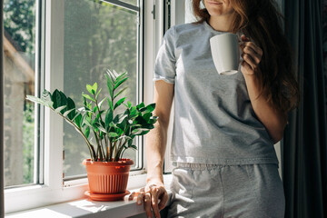 A woman in pajamas drinks coffee and stands by the window with a mosquito net. There is a potted...