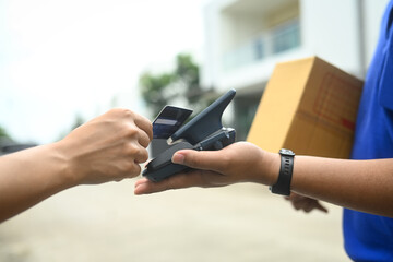 Unrecognizable man making contactless payment with a credit card while receiving parcel box from delivery man