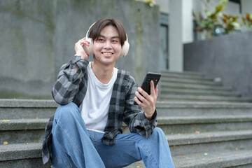 Obraz premium Happy young Asian student wearing wireless headphones and smiling happily while looking at mobile phone at university, college, field trip Learning concept, success.