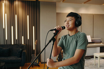 Asian teenager singing into microphone in recording studio with acoustic panels and various musical equipment present in background