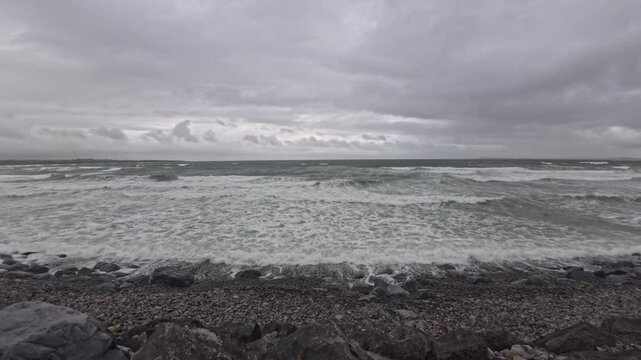Dramatic waves crash against the rocky shore of Strandhill Beach, Ireland, under a stormy sky. The turbulent sea, stirred by strong winds, creates an intense and dynamic coastal scene