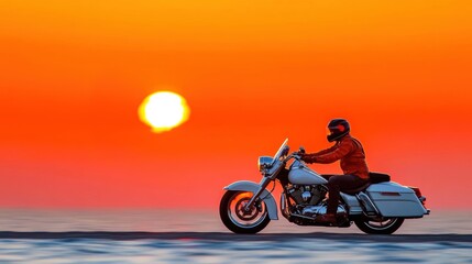 A motorcyclist cruises down a coastal road as the sun sets, casting a warm glow over the landscape and reflecting in the water