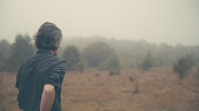 Young man of European appearance in a black shirt with long black hair in nature, fog, morning, autumn, looking at phone, hides it in back pocket, looks around, medium shot, Easter shot