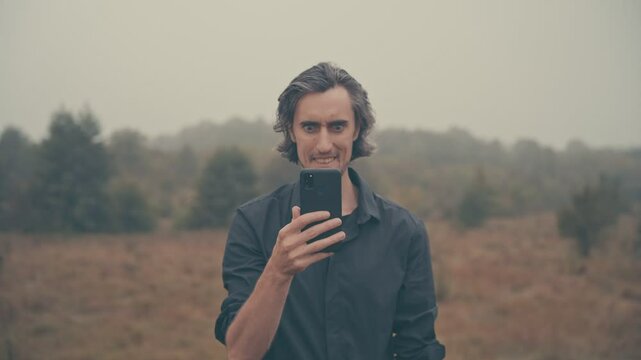 Young man of European appearance in a black shirt with long black hair in nature, fog, morning, autumn, looking tensely at phone, in front of face, smiling exaggeratedly, crazy about social media
