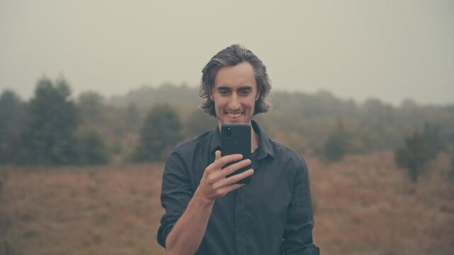 Young man of European appearance in a black shirt with long black hair in nature, fog, morning, autumn, looking crazily at phone, in front of face, smiling exaggeratedly,crazy about social media