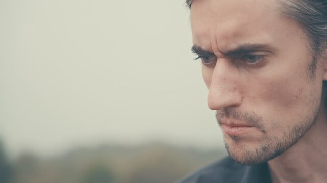 Young man of European appearance in a black shirt with long black hair, unshaven, profile, looking down seriously, turns gaze to camera, tenses jaw, close-up.
