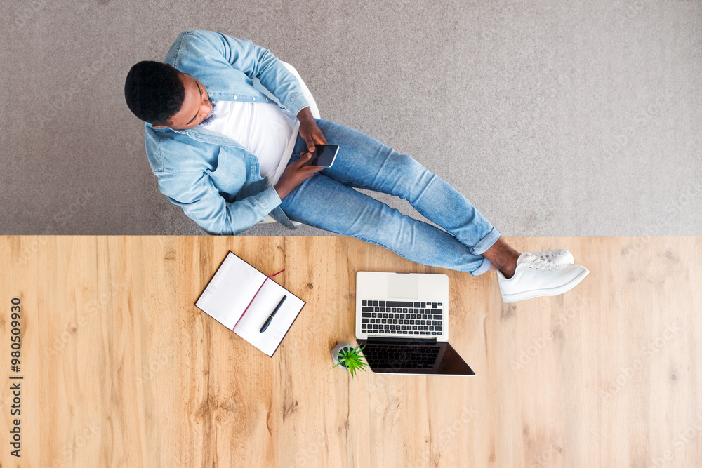 Poster idleness at work. top view of carefree afro employee using smartphone and keeping his legs on table 