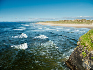 Green grass grows on cliff and amazing waves and sandy Bundoran beach in Ireland on warm sunny day. Popular tourist and surfers area. Blue cloudy sky. Irish landscape. Rich saturated color.