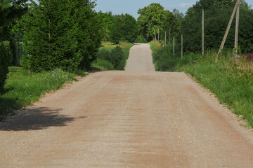 A rural straight dirt road with dip and trees in the background. The road is empty and there is no one on it. Warm sunny day. Country side commute and transportation.