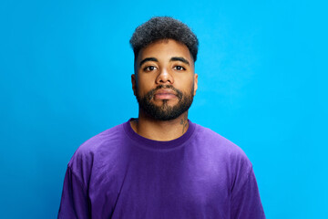 Bearded African-American man in purple shirt stands in relaxed pose, looking directly at camera against blue background. Concept of human emotions, youth, social media, fashion and style.