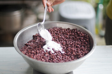 Pouring tempeh or tempe starter (Rhizopus oligosporus,  Rhizopus oryzae) to dried boiled azuki (adzuki) in mixing bowl on white table closed up selective focus blurred background