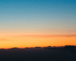 Sunrise and waning moon above black clouds and below clear sky with beautiful colors from blue to orange