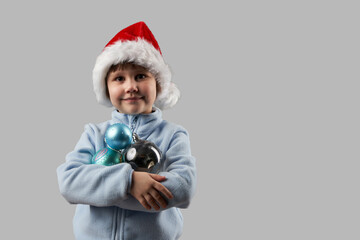 Child in Santa Claus hat with Christmas tree toys (glass balls) for Christmas tree decoration in hands on gray background. Copy space.