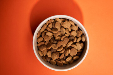 A top-down view of a bowl filled with dog food placed on an orange background. The close-up shot emphasizes the texture and color contrast.