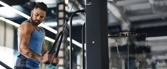 A man focuses intently while performing a cable rope workout in a well-lit gym, panorama with copy space