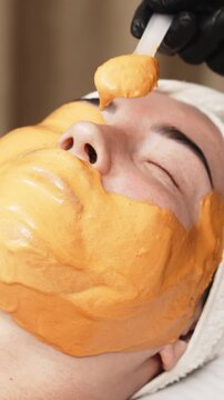 Close-up of a cosmetologist applying an alginate mask to a client's face with a spatula. The scene highlights professional facial skin care treatments in a beauty salon.